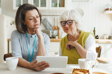 Female volunteer of retirement home nursing her senior female patient, watching series together or...