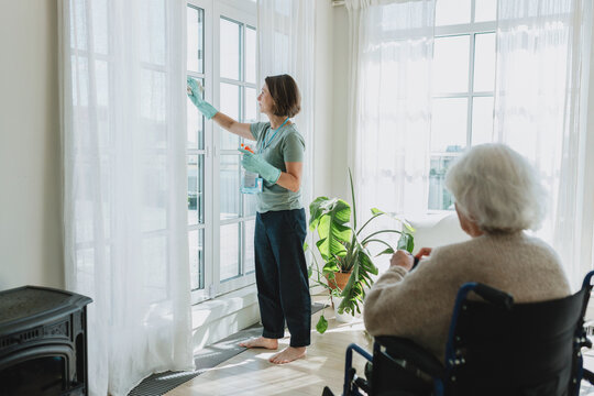 Cleaning service for seniors concept. Female cleaner in gloves washing big panoramic windows in cozy living-room using detergent and talking to old disabled lady home owner in wheelchair