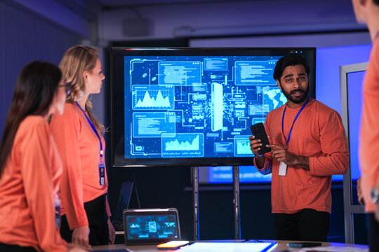 In a government command center, a diverse team of intelligence analysts collaborates on a critical mission, analyzing real-time data from a tablet, phone, and a large video wall.