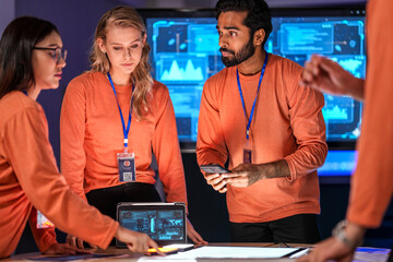 A diverse tech event team in matching orange uniforms collaborates in a high-tech control room, coordinating logistics by analyzing real-time data on a tablet and smartphone.