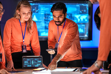 A diverse tech event team in matching orange uniforms collaborates in a high-tech control room, coordinating logistics by analyzing real-time data on a tablet and smartphone.
