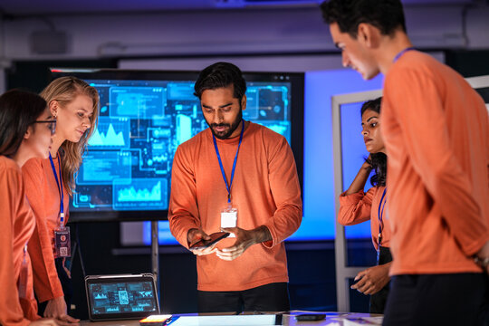 In a government command center, a diverse team of intelligence analysts collaborates on a critical mission, analyzing real-time data from a tablet, phone, and a large video wall.