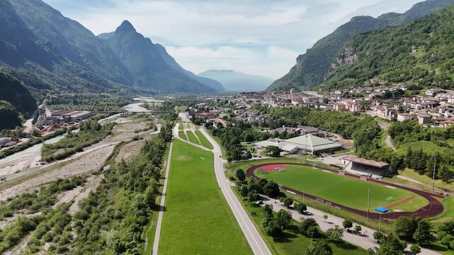Drone flight over Longarone valley &mdash; green slopes, alpine river, soccer field, and the road cutting through the heart of this sunny Dolomites town.