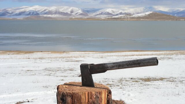 An axe is left in a tree stump, a vital tool for chopping firewood in the harsh Mongolian winter. In the background, a lake and snow-covered mountains complete the scene.