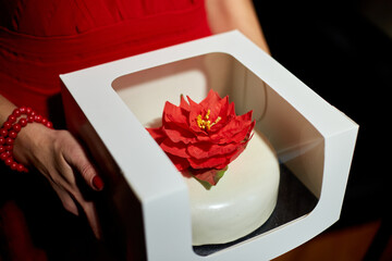 A woman wearing a striking red dress presents an elegant cake adorned with a red flower, showcasing meticulous craftsmanship. The festive atmosphere enhances the moment.