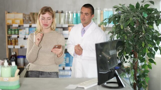Woman scrolls through online catalog on the pharmacy website against the background of a pharmacist. Customer holds a mobile phone in hands in a pharmacy and looking at shelves with goods. High