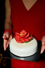 The woman presents an elegantly crafted cake featuring a vibrant flower on top. The surroundings are softly lit, enhancing the festive atmosphere of the occasion.