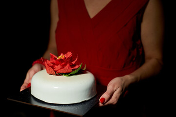 The woman presents an elegantly crafted cake featuring a vibrant flower on top. The surroundings are softly lit, enhancing the festive atmosphere of the occasion.