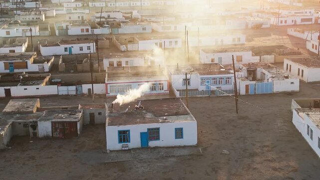 Aerial view of a small village along the Pamir Highway, Tajikistan. White flat-roof houses with smoke rising under golden sunset light on the high-altitude plateau