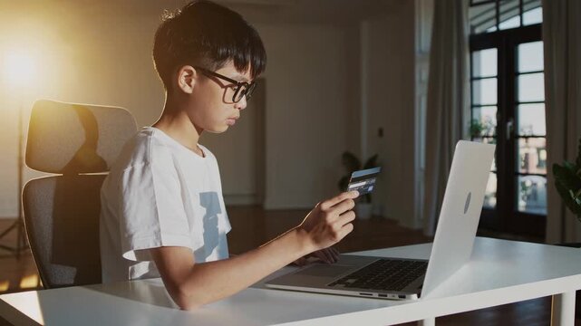 Asian boy engages in online shopping, holding credit card while seated at a desk with laptop, demonstrating modern payment methods, camera follows action smoothly