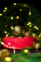 A vibrant red cake sits elegantly on a dark surface, decorated with golden orbs and festive greenery, creating a joyful atmosphere with twinkling holiday lights.