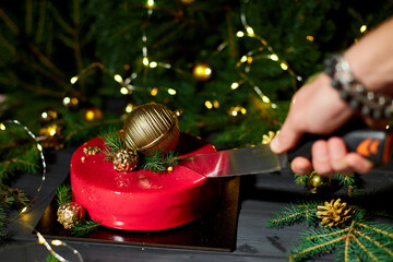A hand holds a knife to slice a vibrant red cake decorated with golden ornaments and pine, surrounded by twinkling lights in a cheerful holiday atmosphere.