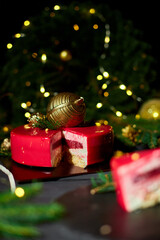 A slice of colorful layered dessert sits on a slate serving plate, surrounded by green pine branches and shimmering holiday decorations, evoking festive cheer.