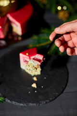 A hand reaches for a slice of colorful layered cake, with a rich red topping, placed on a dark plate surrounded by soft, twinkling lights and holiday decorations.