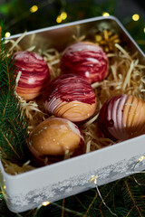 An elegant assortment of colorful dessert spheres is beautifully arranged in a festive gift box filled with straw and surrounded by fresh greenery and decorative touches, perfect for celebrations.