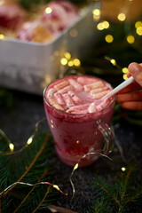 A delicious holiday dessert drink features a rich pink color, topped with fluffy marshmallows, as it is held against a backdrop of festive decorations and soft lights.