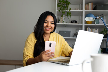 Smiling latin american woman using laptop and smartphone at home office