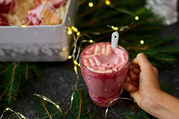A delicious holiday dessert drink features a rich pink color, topped with fluffy marshmallows, as it is held against a backdrop of festive decorations and soft lights.