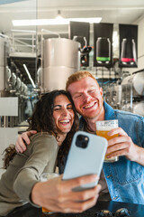 Couple smiling and taking a picture together with a smartphone while visiting a modern brewery and tasting craft beer