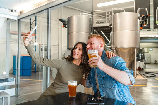 Smiling young couple having fun, enjoying craft beer and taking selfie during brewery tour in modern industry facility - Powered by Adobe