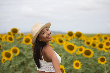 A beautiful black Latin woman wearing a white top and a straw hat stands among sunflowers. The...