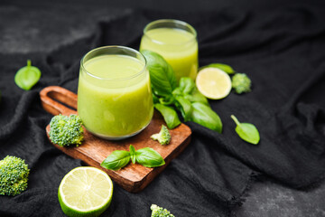 Bright green smoothie in glasses with fresh broccoli and lime wedges on wooden board and black cloth.