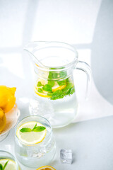 Close up of jug filled with lemon water mint leaves and ice cubes placed on light surface in sunlight