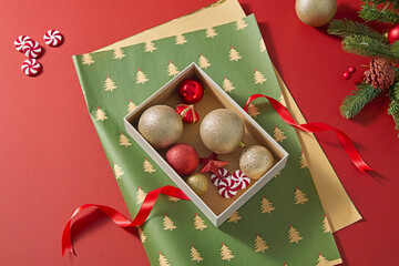 Open Christmas gift box filled with ornaments, green wrapping paper, and red ribbon on red table. Cozy holiday concept for background or seasonal promotion.