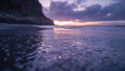 Glistening tide-marked shore with soft surf approaching stone bluff beneath moody overcast at dusk glow