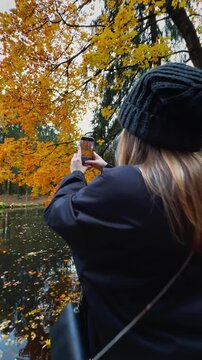 Back view, full shot of a woman in a black outfit and knit hat standing on a massive tree stump by a pond, taking a vertical photo of the vibrant golden autumn foliage and forest reflections on a clou