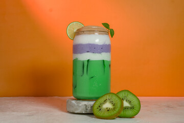 Layered kiwi and taro iced drink in a glass jar with lime slice and mint leaves. Refreshing colorful beverage with gradient layers and bright orange background.