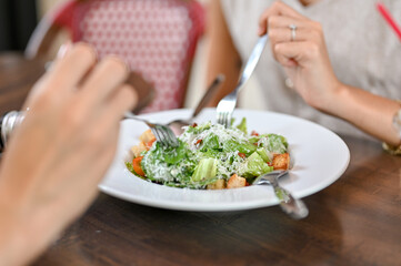 Close up woman's hands holding fork eating salad with friend sits at wooden table in restaurant cafe