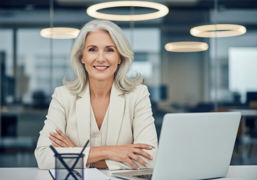 Elegant senior businesswoman smiling with arms crossed at her desk with laptop image photo