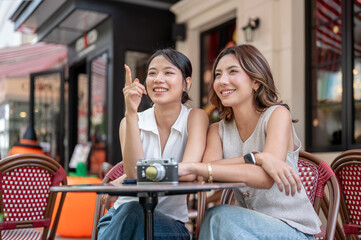 Pretty asian woman pointing beside friend while sitting with camera at round table outside the cafe