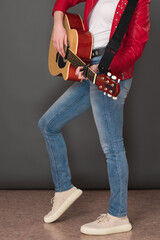 Side view of lower half of an unrecognizable female guitarist playing six string acoustic guitar. Stylish youthful musician is wearing in red leather jacket, blue jeans and sneakers
