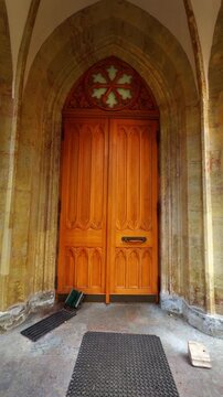 Symmetrical, vertical full shot of the striking, bright wooden double doors of a historical church, set within a weathered stone gothic archway with a quatrefoil window above and dark rubber mats on