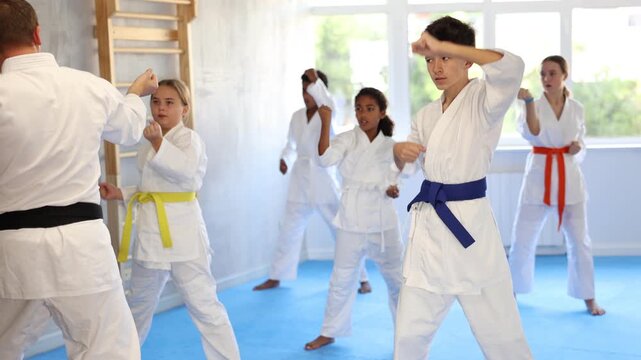 Determined boy in white kimono practicing punches in gym during martial arts workout with multiracial group of tweens. Shadow fight, combat sports training concept 