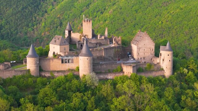 bourscheid castle ch&acirc;teau de green cover hills walls towers flags parked visitor vehicles old historical site to left near right drone panning shot 