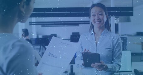 Smiling adult Asian woman holding tablet in open-plan office, with printed chart and monitors