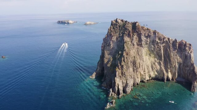 Aerial view of the dramatic Basiluzzo Islet, its rocky texture contrasting with the deep blue sea and a boat leaving a white trail, Panarea, Sicily, Italy.