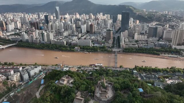 Aerial view of the city of Lanzhou with a muddy Yellow River flowing through, contrasting with the dense urban landscape and verdant hills, Lanzhou, Gansu, China.