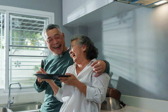 Happy Asian senior couple laughing and playfully teasing each other in kitchen during morning, showing love and joyful retirement life. - Powered by Adobe