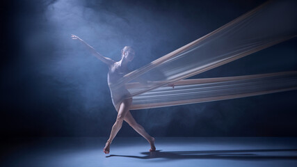 Ballet dancer crossing legs while wrapped in flowing fabric on dark stage. Concept of performance movement, body alignment, and contemporary choreography in studio lighting.