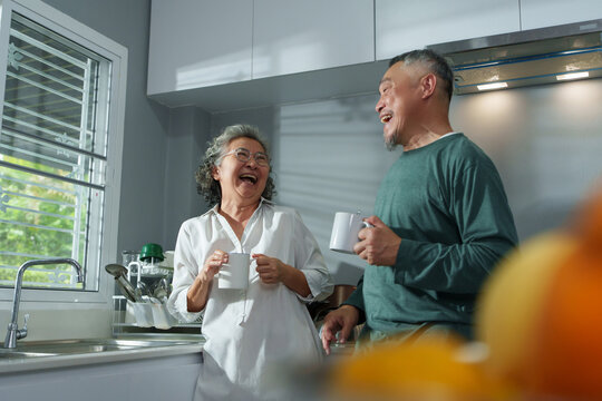 Smiling Asian senior couple drinking coffee and laughing together in kitchen, showing love and happiness in retirement life.