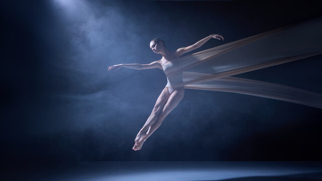 Ballet dancer leaping with fabric tied around body under blue light. Concept of movement energy, performance art, and dance flexibility for editorial and advertising use.