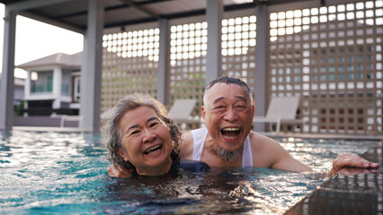 Happy Asian senior couple smiling at camera in swimming pool at sunset, showing love, joy, and peaceful retirement lifestyle together.