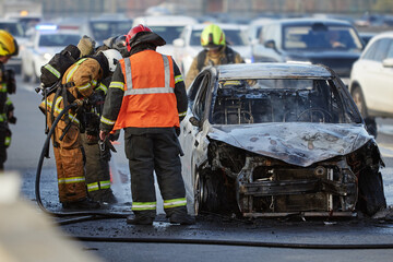 Group of firefighters and emergency responders extinguishing burning car on city street, wearing...