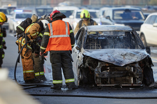 Group of firefighters and emergency responders extinguishing burning car on city street, wearing protective gear and helmets, smoke rising from damaged vehicle, traffic visible in background