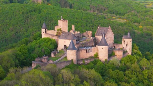 vast area bourscheid castle ch&acirc;teau de green valley hills behind walls black domed towers flag top foggy day tall trees colorful to left around right drone panning 