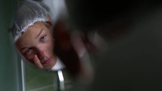 A girl looks at herself in a round mirror in a hotel bathroom and adjusts the clear shower cap on her hair. The teenager is performing her morning or evening body care routine.